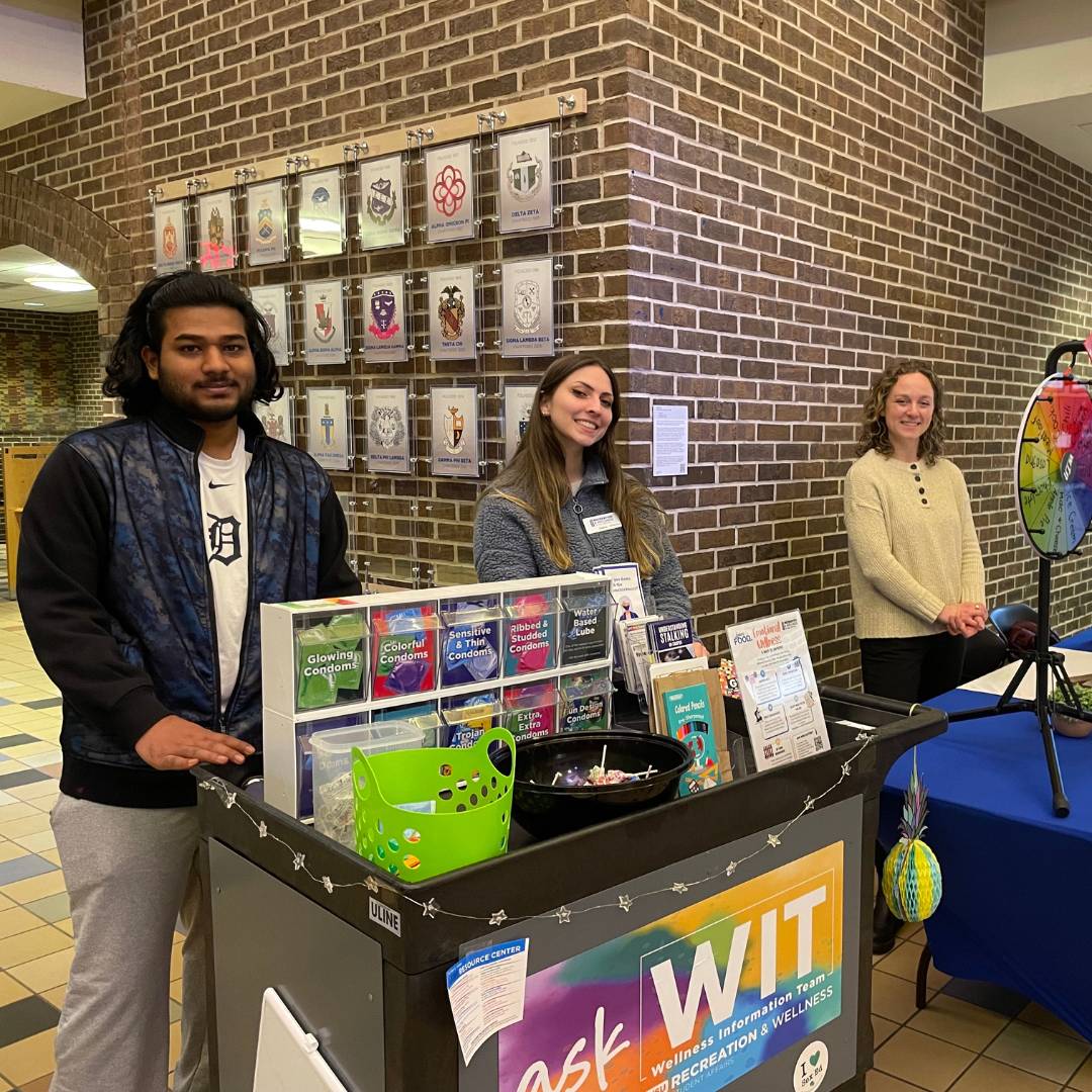 Image of two WIT Peer Educators tabling in Kirkhof behind the WIT cart and the on-campus registered dietitian behind a spinning wheel